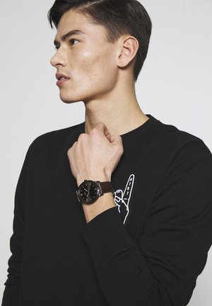 Jeune homme en chemise noire à manches longues avec détail graphique tenant son poignet, portant une montre noire avec un bracelet en cuir marron, faisant face à gauche.