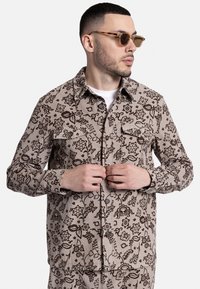 Μπλούζα κοντομάνικη από καστανό υλικό με μπεζ βάση και σκούρο λουλουδάτο μοτίβο, διαθέτει κουμπιά μπροστά και δύο τσέπες στο στήθος.