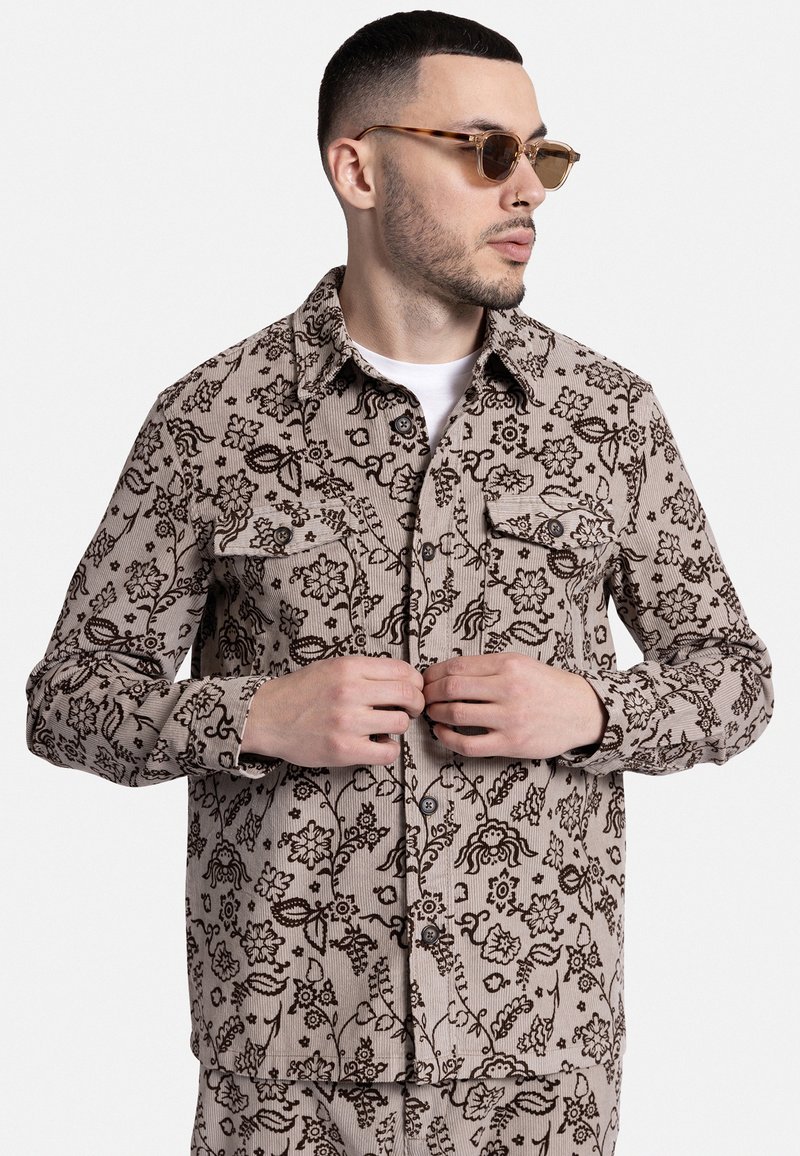 Μπλούζα κοντομάνικη από καστανό υλικό με μπεζ βάση και σκούρο λουλουδάτο μοτίβο, διαθέτει κουμπιά μπροστά και δύο τσέπες στο στήθος.