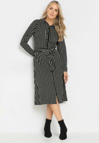 Schwarz-weiß gestreiftes Hemdkleid mit langen Ärmeln, durchgehender Knopfleiste, tailliertem Gürtel und einem Schlitz bis zum Oberschenkel. Kombiniert mit schwarzen Knöchelstiefeln.