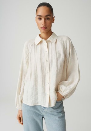 Frau mit nach hinten gebundenen Haaren, die eine lockere, langärmelige, cremefarbene Bluse mit Knöpfen und hellblaue Jeans vor einem schlichten Hintergrund trägt.