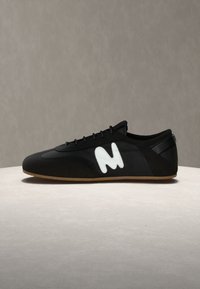 Μαύρο αθλητικό παπούτσι χαμηλού προφίλ με κορδόνια, λευκό στυλιζαρισμένο λογότυπο "M" στην πλευρά και ανοιχτό καφέ σόλα, παρουσιασμένο σε ουδέτερο φόντο.