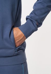 Mornarska jopica z gladko teksturo, ki ima žep z zadrgo. Manšete imajo majhen, kontrastni logotip.