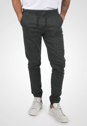 Pantalones de chándal negros con cinturilla elástica, bolsillos laterales, puños ajustados y costuras en contraste; combinados con zapatillas blancas.