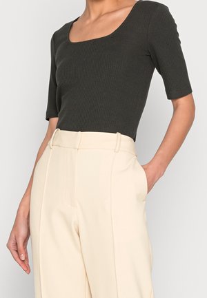 Vrouw draagt een donkergroene geribbelde korte mouwen top, ingestopt in een crème kleurige, brede broek met hoge taille, hand in de zak.