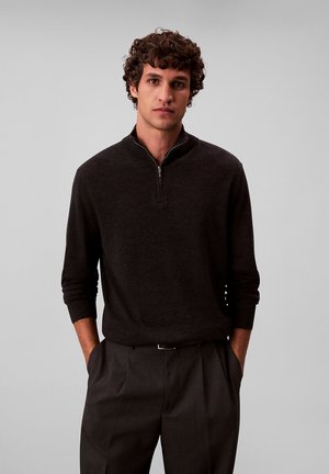 Giovane uomo con capelli ricci che indossa un maglione scuro a maniche lunghe con mezza cerniera e pantaloni scuri, in piedi con le mani in tasche.