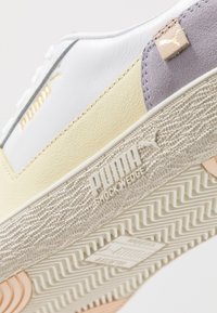 Sneakers Puma z białą i żółtą skórzaną cholewką, purpurowymi akcentami z zamszu, teksturowaną podeszwą oraz wytłoczonym logo na pięcie.