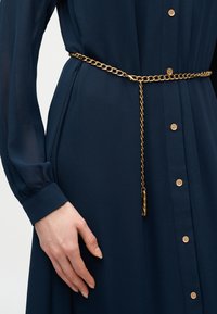 Robe boutonnée bleu marine avec manches longues transparentes, cintrée à la taille par une fine ceinture chaîne dorée à bout pendante.