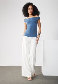 Top azul ajustado y fuera de los hombros con una textura suave, combinado con pantalones blancos sueltos y sandalias plateadas sobre una superficie gris.