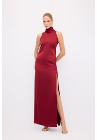 Rød satin maxi kjole med høj hals, skulderbærende design og sideslids; glat tekstur og tætsiddende silhuet. Guld hælede sandaler.