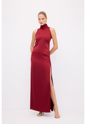Rød satin maxi kjole med høj hals, skulderbærende design og sideslids; glat tekstur og tætsiddende silhuet. Guld hælede sandaler.