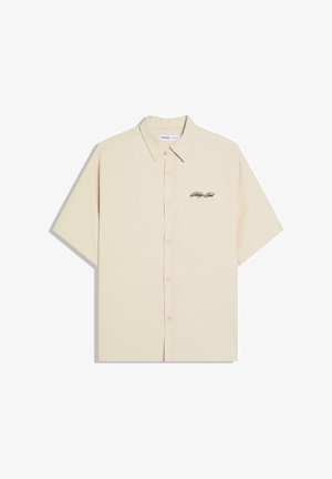 Beżowa krótka koszula na guziki z kołnierzykiem i małym czarnym logo w formie napisu na lewej piersi.