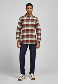 Geruite flanellen shirt in rood, beige en bruin; lange mouwen met knoopmanchetten; gecombineerd met donkerblauwe jeans en witte sneakers met bruine accenten.