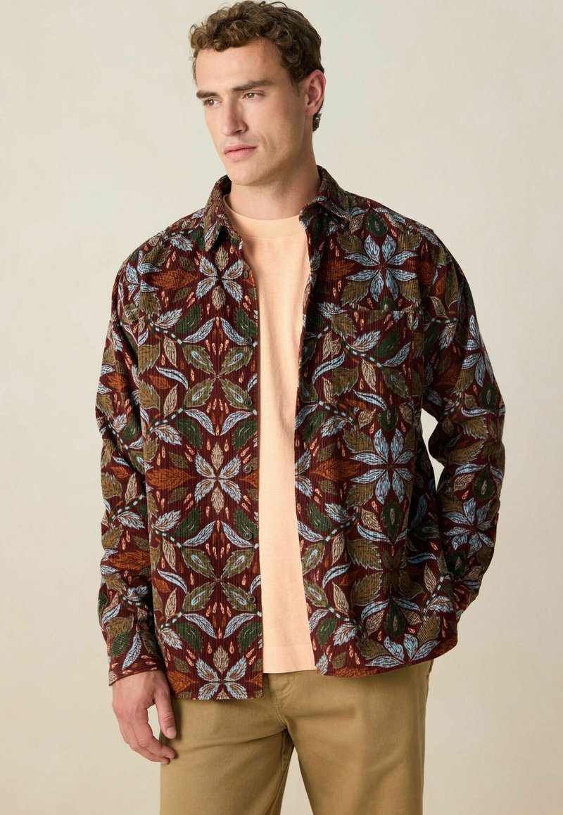 Long-sleeve button-up shirt in brown with floral print featuring blue, green, and rust accents. Worn over a light peach t-shirt.