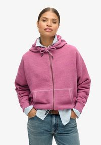 Sweatshirt rose à capuche avec une fermeture éclair, un cordon de serrage au cou et des poches avant. Porté par-dessus une chemise à boutons bleu clair et associé à un jean.
