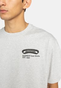 Šedé bavlněné tričko s krátkým rukávem, na kterém je na levé straně hrudi černý grafický potisk "ELEMENT Iron Works EST. 1992".