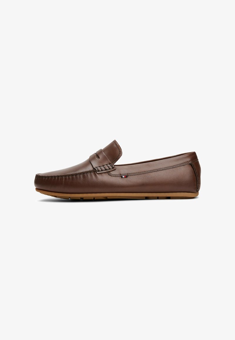 Mocasines de cuero marrón con suela de goma plana, que presentan un detalle de costura y un pequeño emblema en el lateral. Textura suave en general.