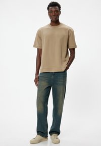 Beige katoenen T-shirt met korte mouwen, gecombineerd met blauwe jeans met een vervaagde textuur en lichtbruine sneakers. Eenvoudig ontwerp, relaxte pasvorm.