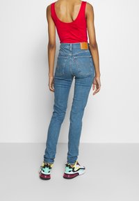 Blue denim high-waisted jeans with a rear logo patch and back pockets, paired with a red tank top and multicolored sneakers.