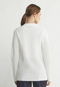 Vit långärmad tröja i meshstickat material, med rund halsringning och en texturerad mönster på ytan.