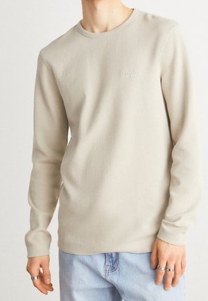 Beige Langarm-Ripp T-Shirt mit Rundhalsausschnitt und dezentem "BOSS"-Logo auf der Brust, kombiniert mit hellblauen Jeans.