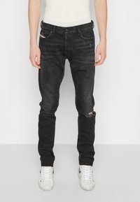 Jeans de mezclilla negra con corte slim, que presentan una rodilla desgastada, un ligero efecto de decoloración y cinco bolsillos. Combinados con zapatillas blancas.