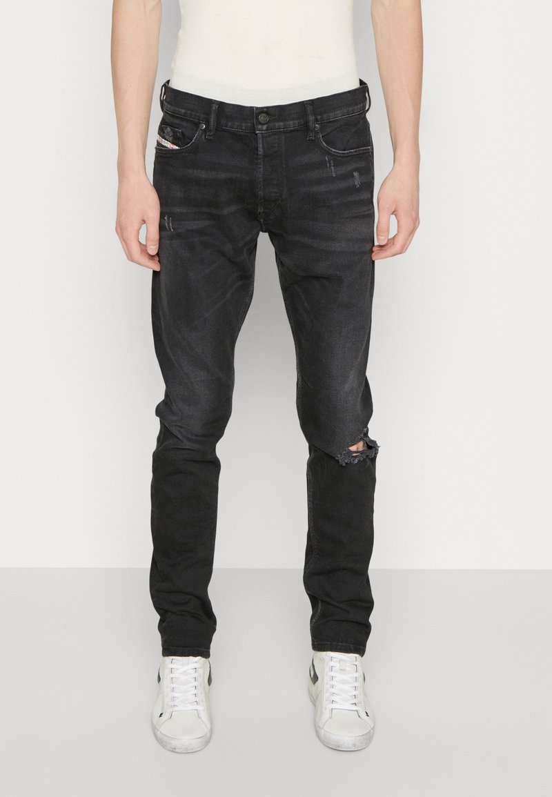 Jeans de mezclilla negra con corte slim, que presentan una rodilla desgastada, un ligero efecto de decoloración y cinco bolsillos. Combinados con zapatillas blancas.