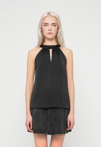 Mulher jovem com cabelo loiro comprido, vestindo um top halter preto sem mangas e uma saia de denim pregueada cinza escuro, de pé contra um fundo simples.