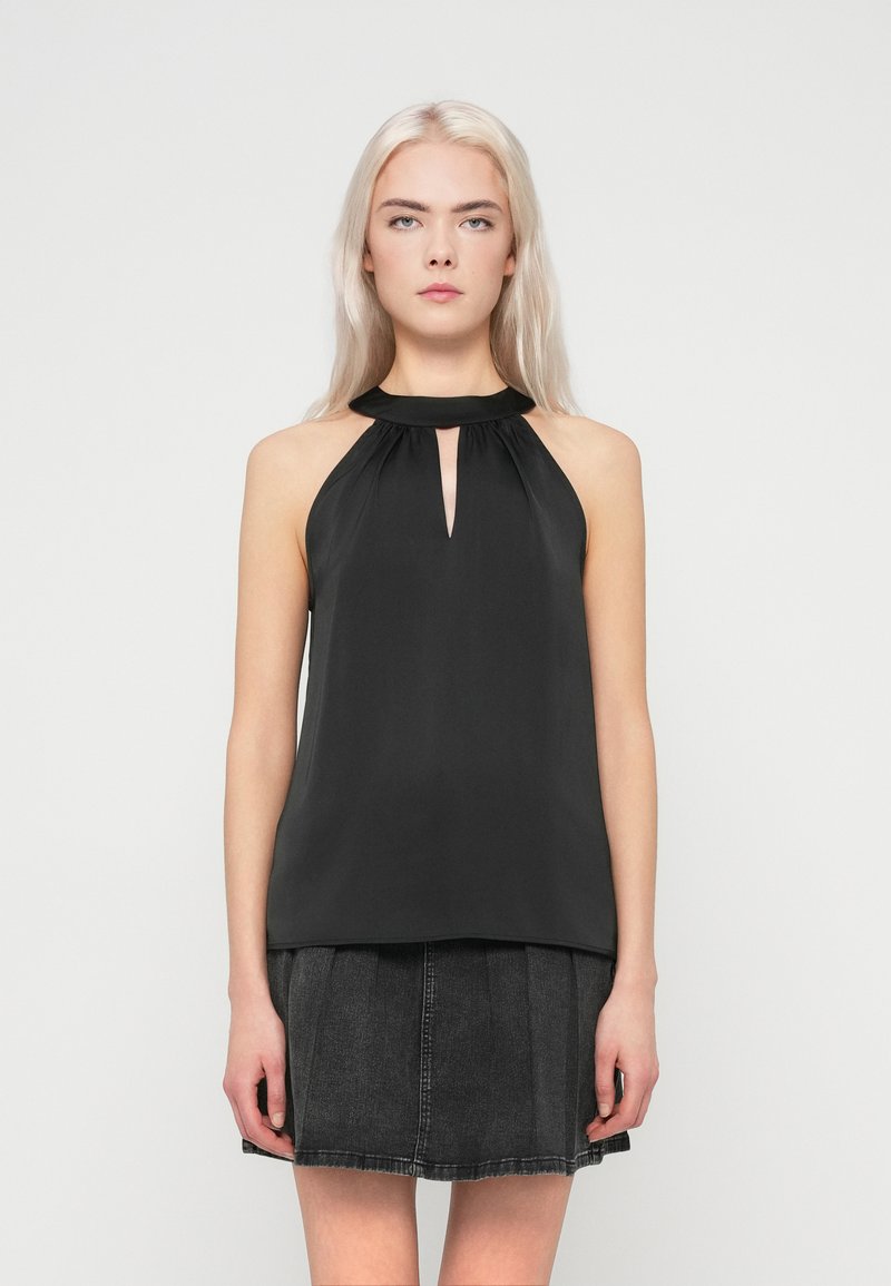 Mulher jovem com cabelo loiro comprido, vestindo um top halter preto sem mangas e uma saia de denim pregueada cinza escuro, de pé contra um fundo simples.