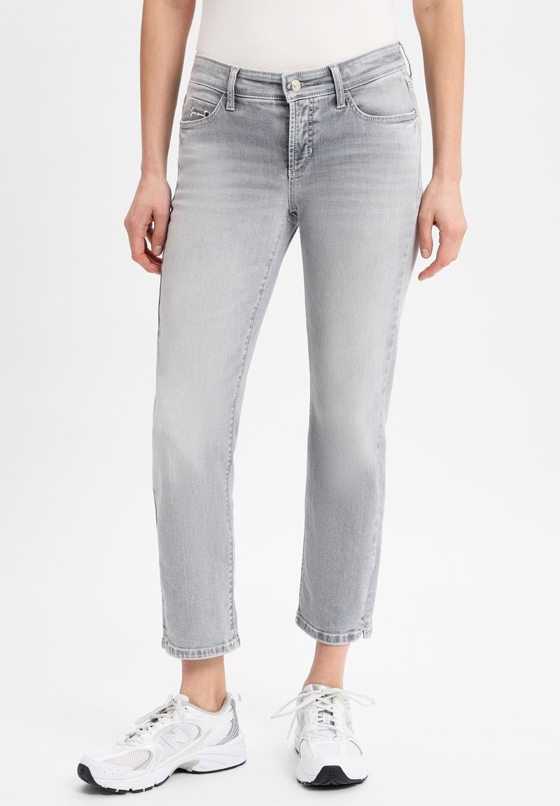 Personne portant un jean gris clair délavé et des chaussures de sport blanches à lacets, debout devant un fond blanc uni.
