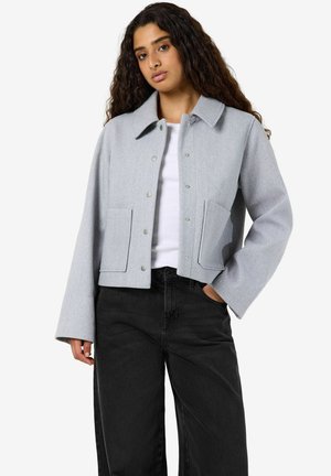 Femme aux longs cheveux bouclés portant une veste boutonnée gris clair sur une chemise blanche et un pantalon noir, une main dans la poche.