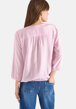 Woman with long brown hair wearing a loose pink and white striped blouse and blue jeans, facing away with one hand in her back pocket.