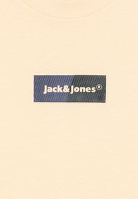 Beige bomulls t-skjorte med et rektangulært blått og mørkeblått logo med "Jack&Jones" i hvit tekst og en teksturert bakgrunn.