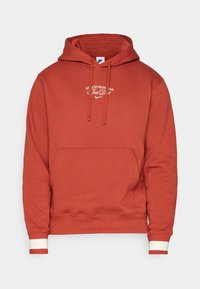 Rostfärgad huvtröja med känguruficka, vita dragsko och kontrasterande ribbade ärmslut. Har "Just Do It"-logotyp i vitt.