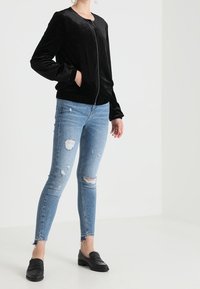 Svart sammet bomberjacka med dragkedja fram, ribbade ärmslut och midja, kombinerad med slitna ljusblå skinny jeans och svarta slip-on skor.