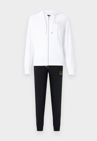 Λευκό φούτερ με κουκούλα και φερμουάρ, με χρυσό λογότυπο, και μαύρο παντελόνι φόρμας με αντίστοιχο χρυσό λογότυπο στο μηρό.