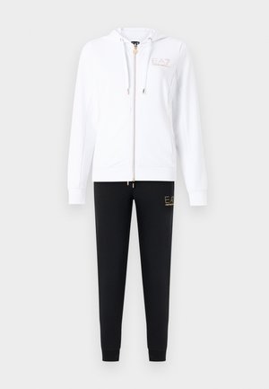 Hvit hettegenser med glidelås og hette, med gullfarget logo, og svarte joggebukser med matchende gulllogo på låret.