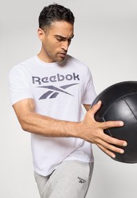 Weißes Kurzarm-Reebok-T-Shirt mit grauem Logo, kombiniert mit hellgrauen Jogginghosen. In einer Hand wird ein strukturierter schwarzer Medizinball gehalten.