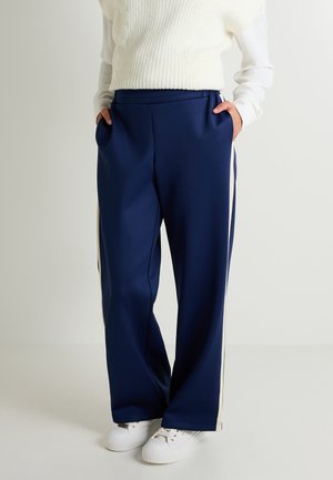 Pantalones de pierna ancha de color azul marino, hechos de una tela suave, con rayas laterales blancas y cintura elástica. Llevados con zapatillas blancas.