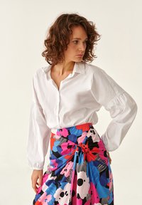 Camisa blanca de manga larga con cierre de botones y mangas fruncidas, combinada con una falda de estampado brillante que presenta diseños florales en azul, rosa y negro.