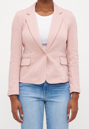 Person wearing a light pink fitted blazer with a single button, white ribbed top, and light blue denim jeans against a plain background.