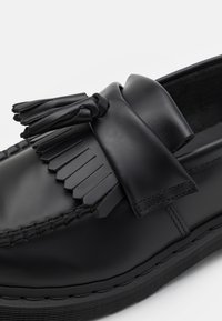 Mocasin de piel negra con detalles de flecos en la parte superior, acentos cosidos y una suela texturizada. Acabado suave con punta redondeada.