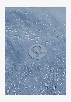 Voda-otporna plava tkanina glatke teksture, s brojnim kapljicama vode i kružnim logotipom u donjem centru.