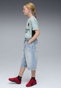 Maglietta sportiva azzurro chiaro con accenti neri, pantaloni di jeans blu e sneakers rosse. La maglietta presenta un logo e un motivo stilizzato.