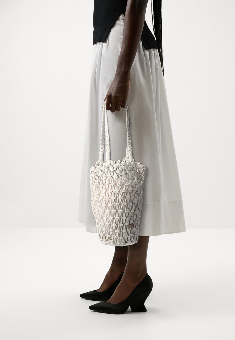 Sac en toile tissé blanc de forme circulaire, doté de poignées tressées et d'un motif texturé. Associé à une longue jupe de couleur claire et des talons noirs.