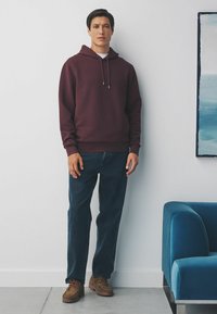 Burgunderfarbener Hoodie mit strukturiertem Material und verstellbaren Kordeln, kombiniert mit dunkelblauen Jeans und braunen Schnürschuhen, steht vor einer weißen Wand.