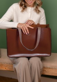 Braune Leder-Tote mit grauen Trägern, rechteckige Form, glatte Oberfläche und Nähdetail, gehalten von einer Person in einem hellen Pullover.