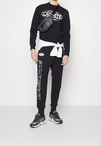 Černá mikina s bílým logem Lacoste, doplněná černými tepláky se stejným logem. Doplňěk: černá crossbody taška s vzorovaným detailem.