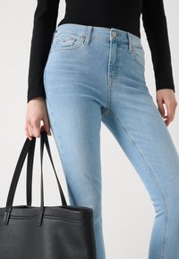 Jean en denim bleu clair avec une coupe slim, doté d'une fermeture à boutons et à zip, et un sac fourre-tout en cuir noir tenu par une main.