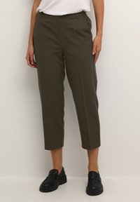 Pantaloni su misura verde oliva con gamba accorciata, pieghe frontali e tasche laterali. Indossati con mocassini neri. Texture liscia e design formale.